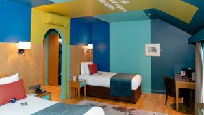Habitación de hotel con cama, mesita de noche, escritorio y silla. Hay una ventana con cortinas, un cuadro en la pared y una puerta que da a otra habitación.