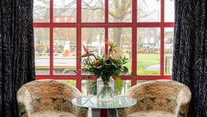 Una habitación con dos sillones con estampado floral frente a una mesa de vidrio con un jarrón de flores. Amplias ventanas con marcos rojos ofrecen vistas al exterior.