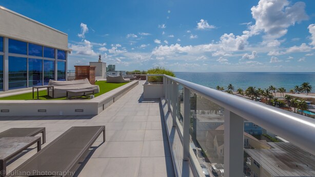 Una terraza en la azotea con mobiliario exterior, vistas al océano y un edificio moderno con grandes ventanales.