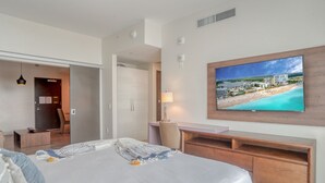 Una habitación de hotel moderna con una cama grande, un televisor montado en la pared mostrando una vista de playa, un escritorio de madera y una puerta que da a otra habitación.