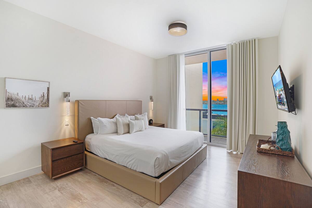 Une chambre moderne avec un grand lit, une table de chevet, un téléviseur à écran plat et une vue sur le coucher de soleil depuis le balcon.