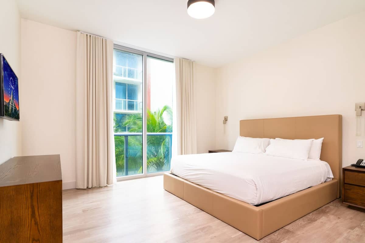 Une chambre d’hôtel avec un grand lit, un bureau en bois, une table de chevet et une vue sur la verdure par la fenêtre.