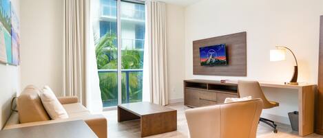 A modern hotel room with a large window, a flat-screen TV, a desk, a chair, and a sofa.