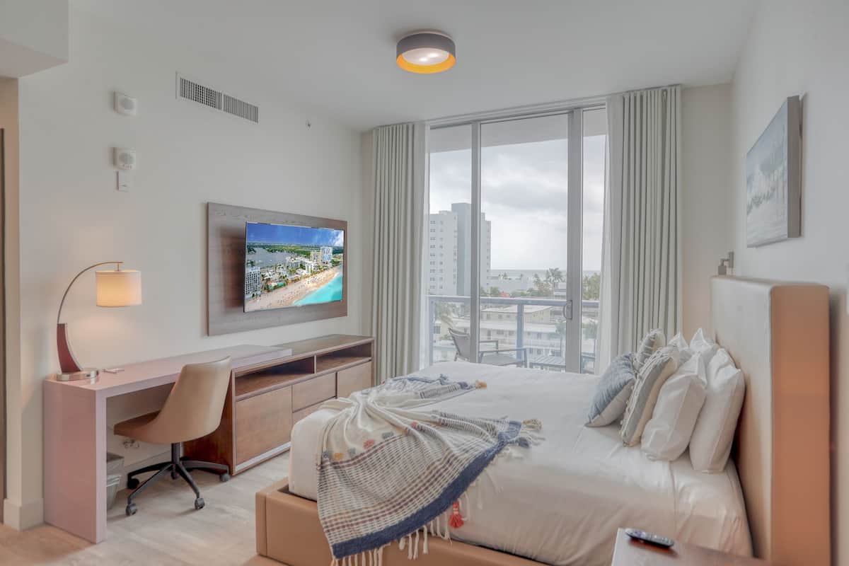 Une chambre d’hôtel équipée d’un lit, d’un bureau, d’une chaise, d’une télévision et de grandes fenêtres donnant sur la ville.