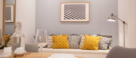 A dining area with a table set for two, a sofa with patterned cushions, a framed abstract artwork on the wall, and a floor lamp.
