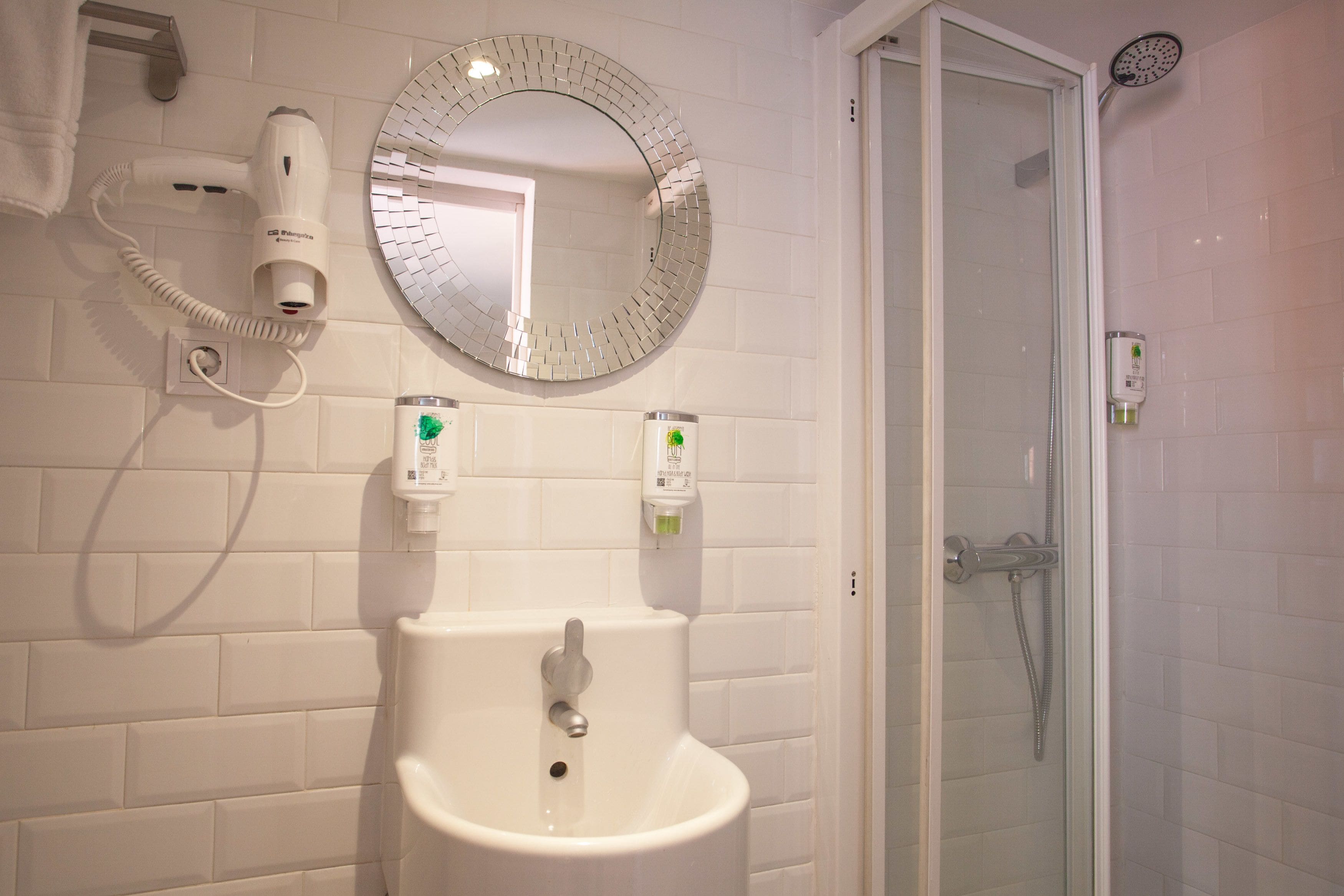 Un baño con un lavabo blanco, un espejo redondo, un secador de pelo y una ducha.