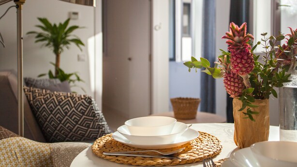 A dining area with a table set for two, featuring a woven placemat, a bowl, a plate, and a fork. There is a potted plant and a lamp in the background. - Gran Central Suites (Madrid)