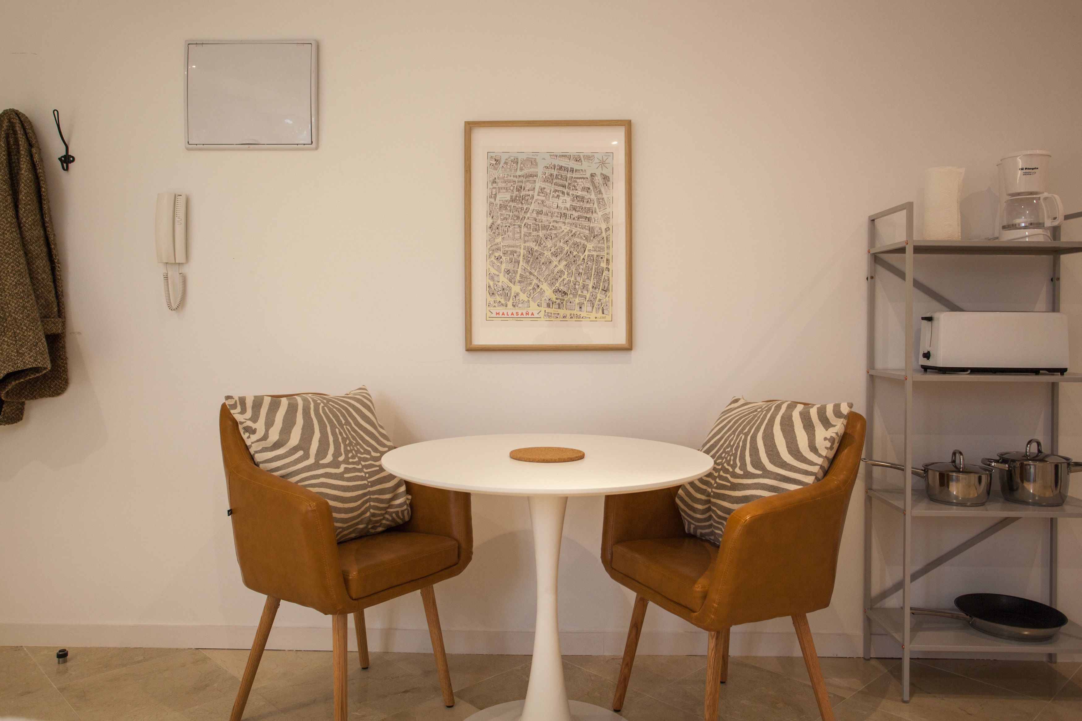 A small, modern room with a round white table, two brown leather chairs with patterned cushions, a framed map on the wall, a telephone, and a metal shelf with kitchen items.