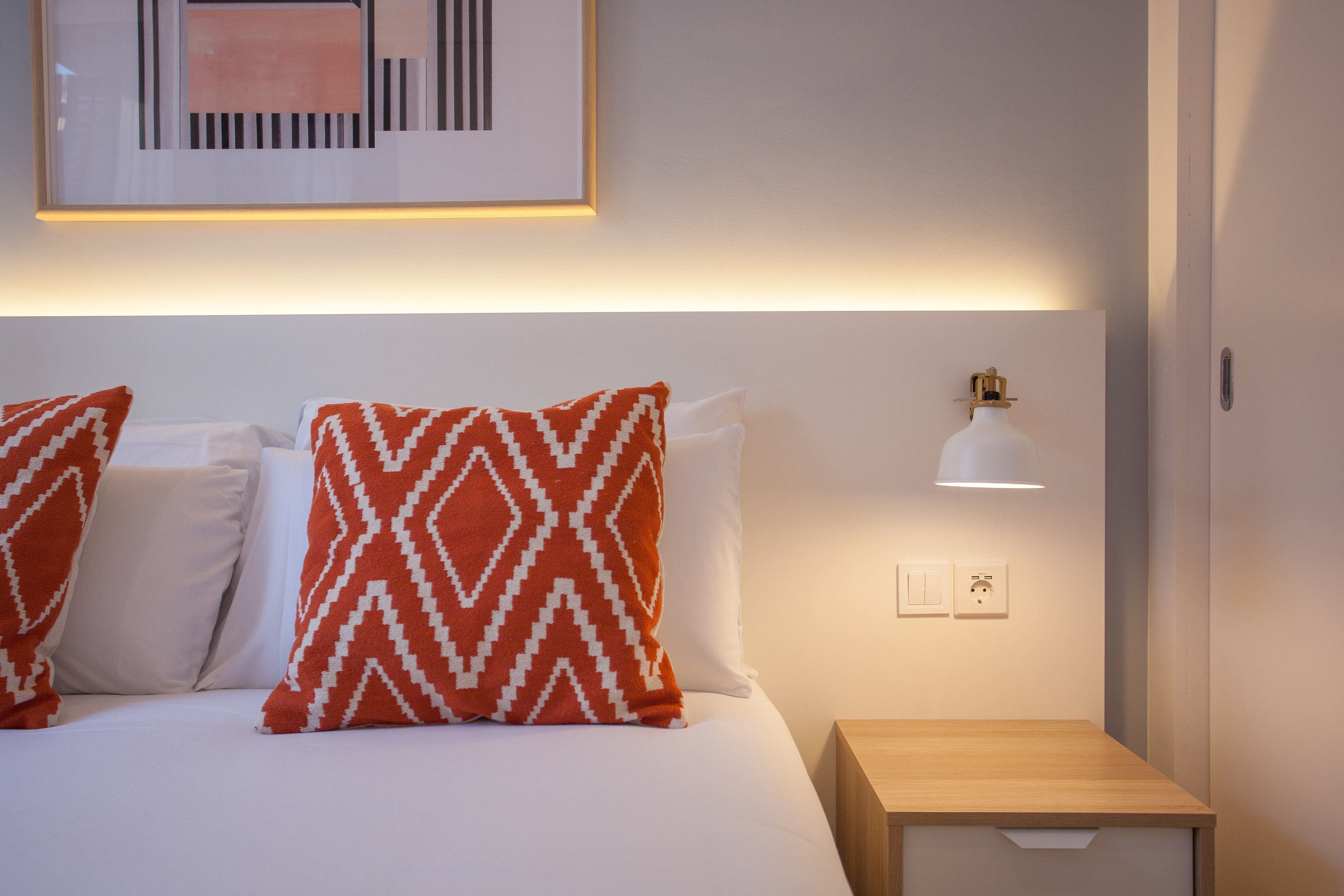 A hotel room with a bed, two patterned pillows, a bedside table, a wall-mounted lamp, and a framed picture above the bed.