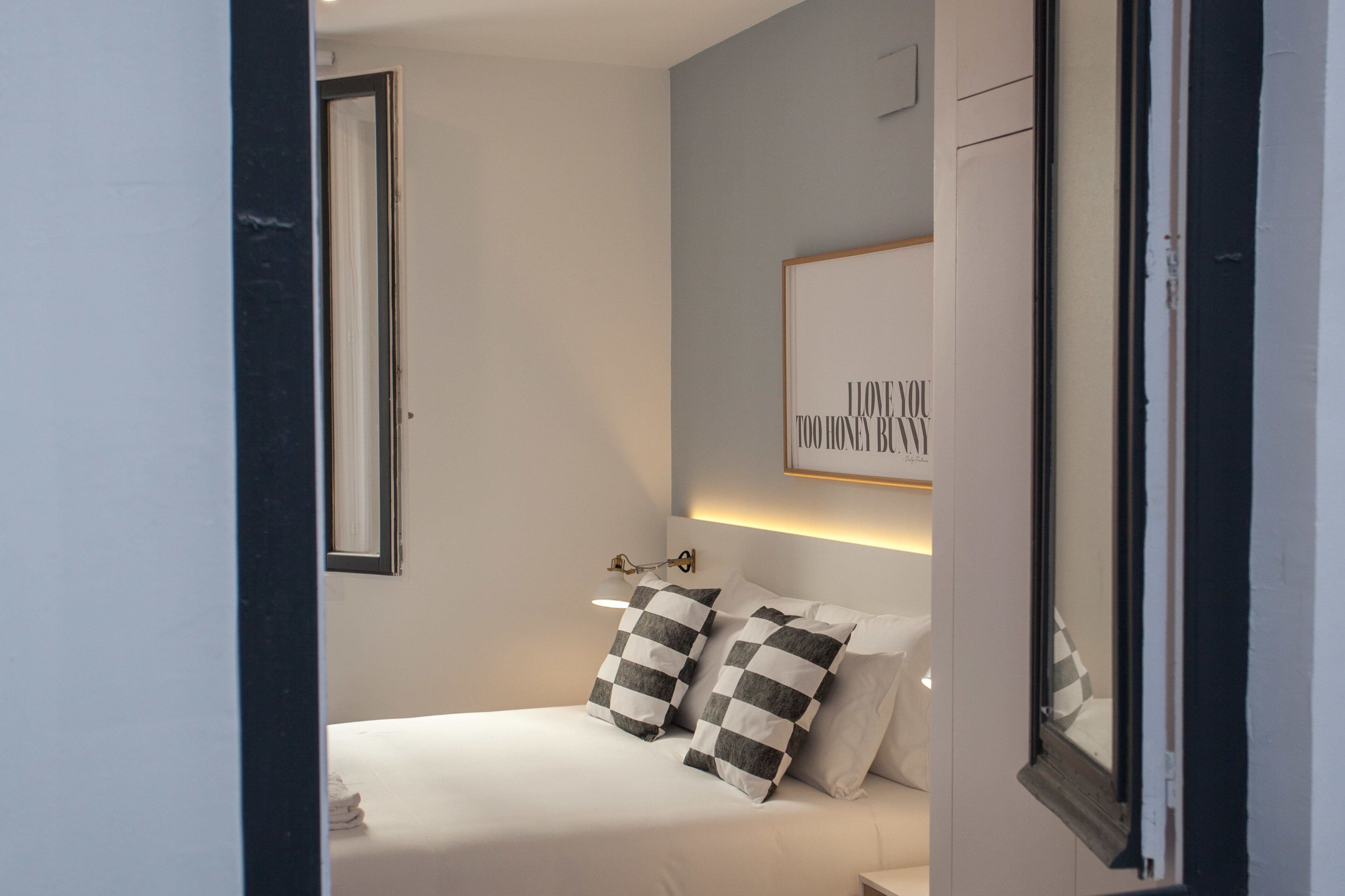 A bedroom with a bed, two pillows, a bedside table, a wall-mounted light, and a framed quote on the wall.