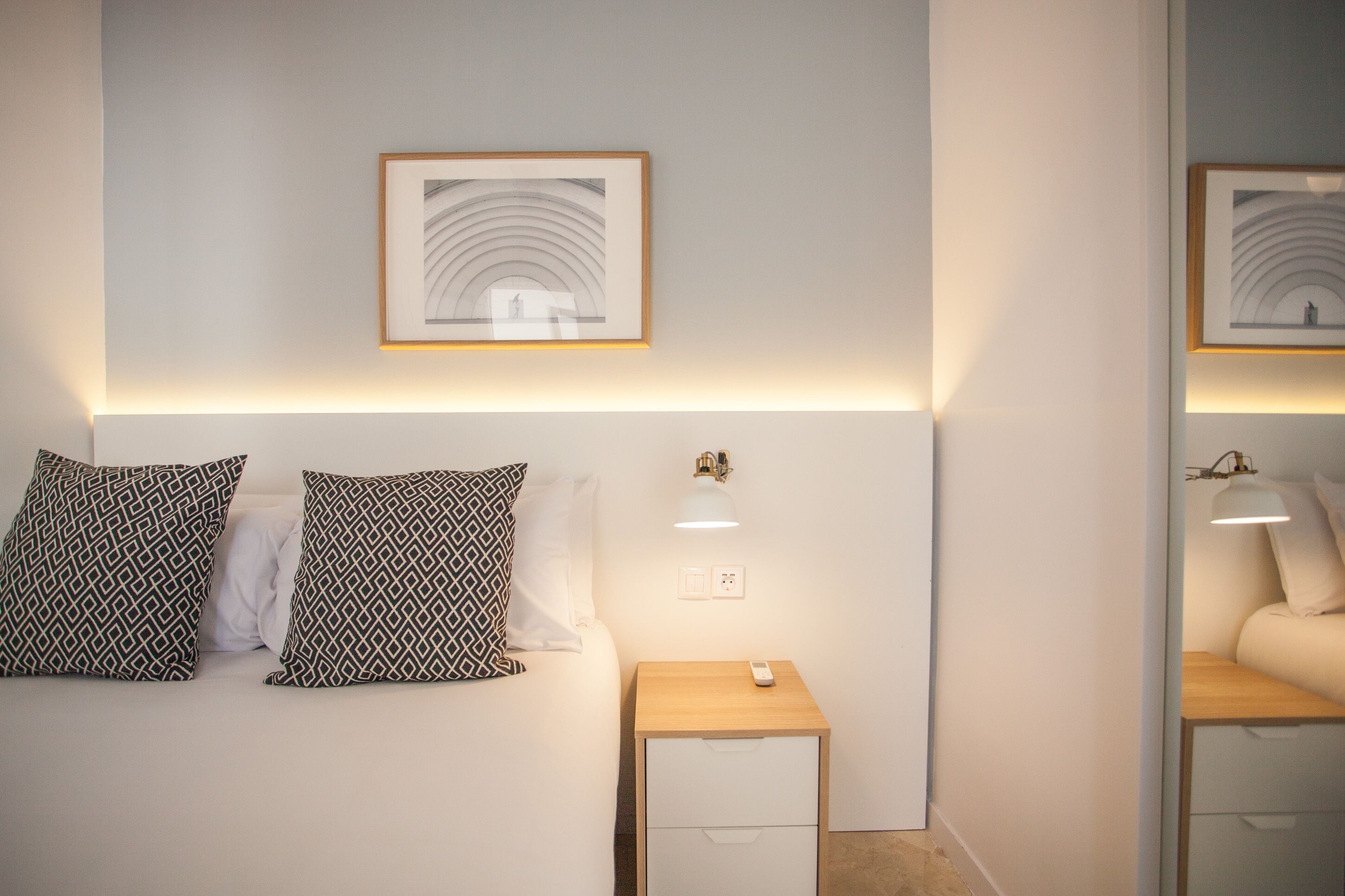 A neatly arranged bedroom with a bed, two patterned pillows, a bedside table with a lamp, and a framed picture on the wall.