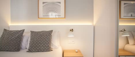 Un dormitorio ordenado con una cama, dos almohadas estampadas, una mesita de noche con una lámpara y un cuadro en la pared.
