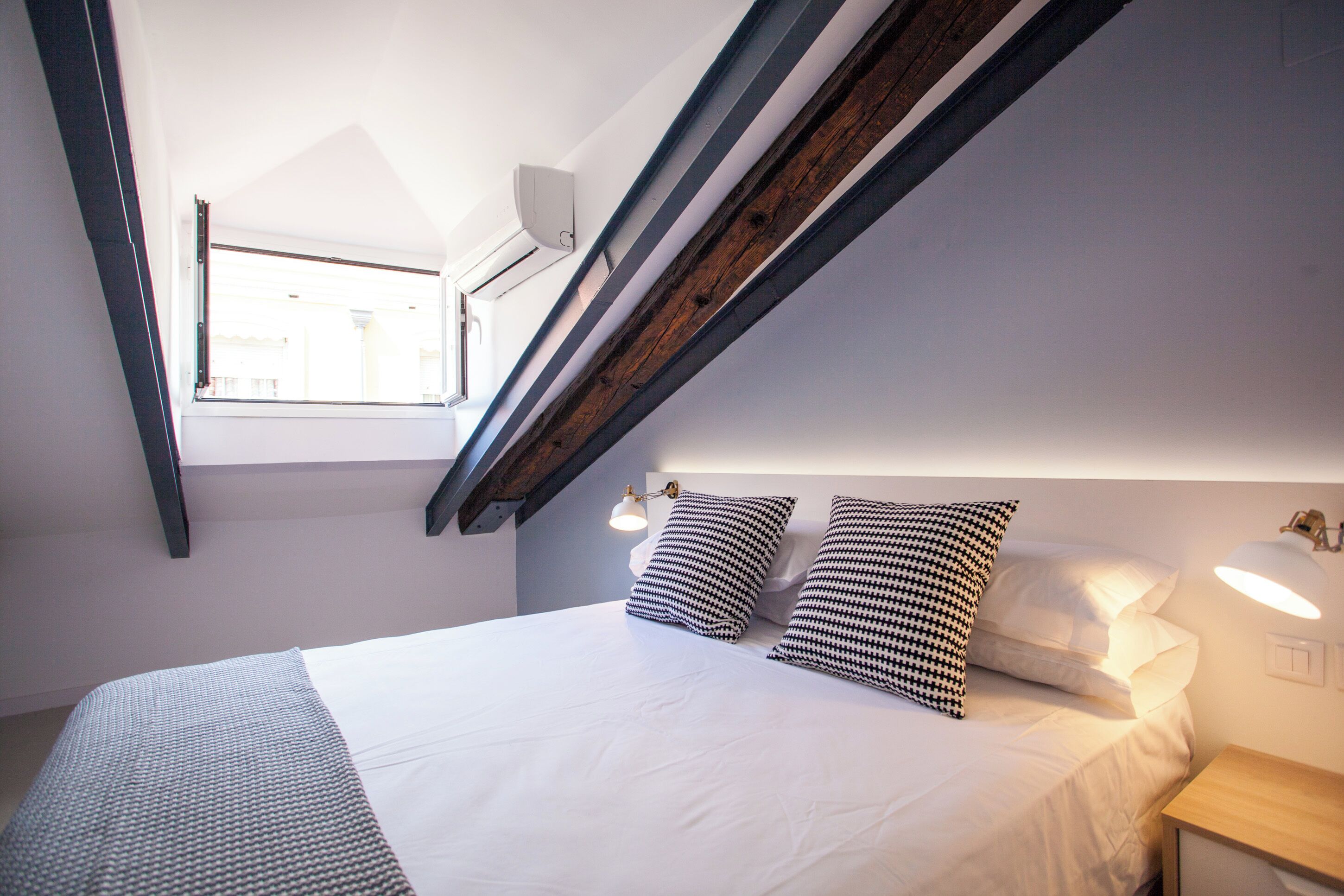 A bedroom with a sloped ceiling, a bed with white bedding, two patterned pillows, a bedside table with a lamp, and a window with a view of buildings.