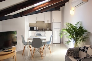 A modern kitchen with white cabinets, a microwave, and a refrigerator. There is a dining area with a table and chairs, a television, and a potted plant. - Gran Central Suites (Madrid)