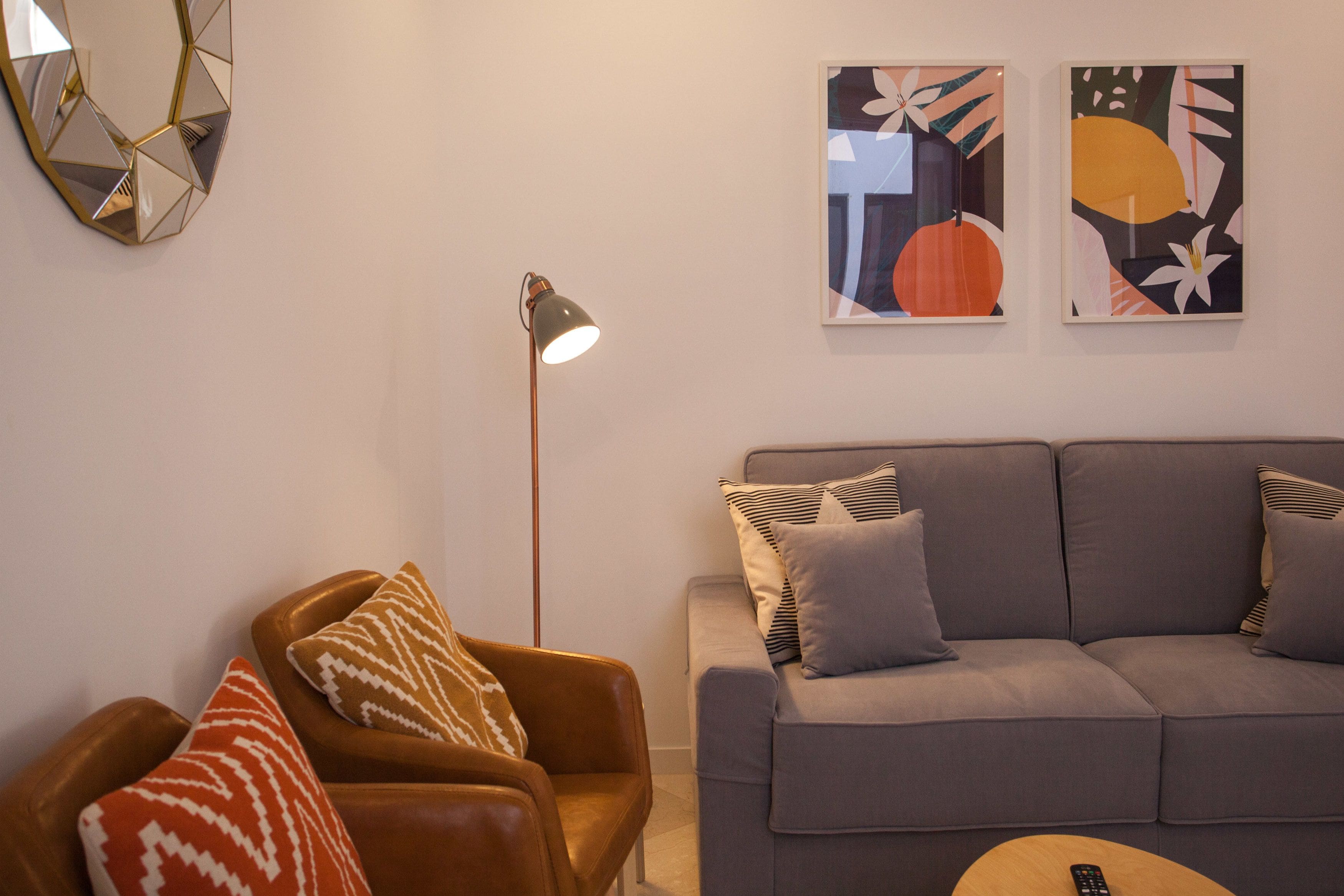 A living room with a brown leather armchair, a grey sofa with patterned cushions, a floor lamp, and two abstract paintings on the wall.