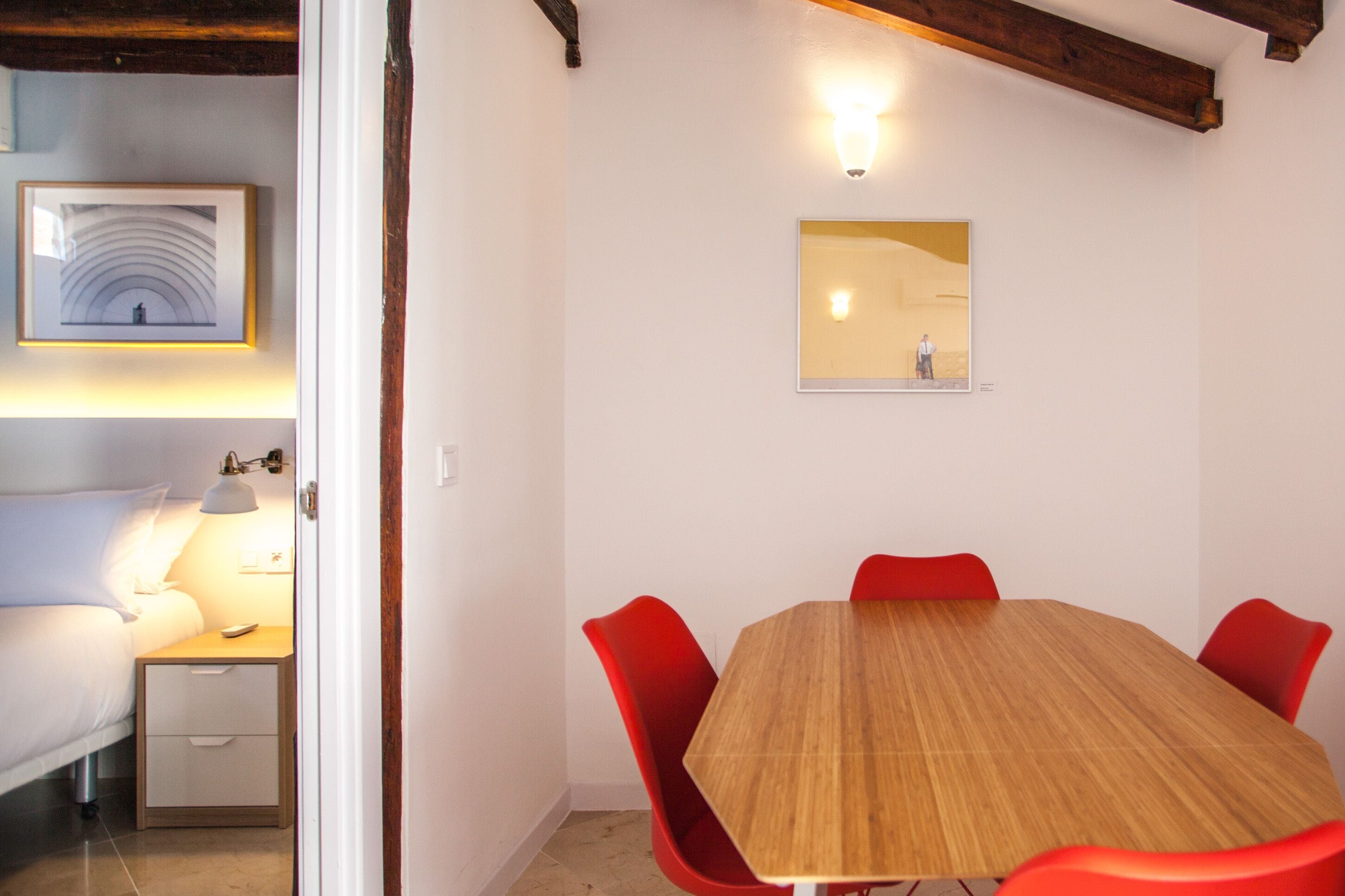 Ein Zimmer mit einem Holzespeisetisch, roten Stühlen, einem Bett mit weißer Bettwäsche, einem Nachttisch mit Lampe und einem gerahmten Kunstwerk an der Wand.
