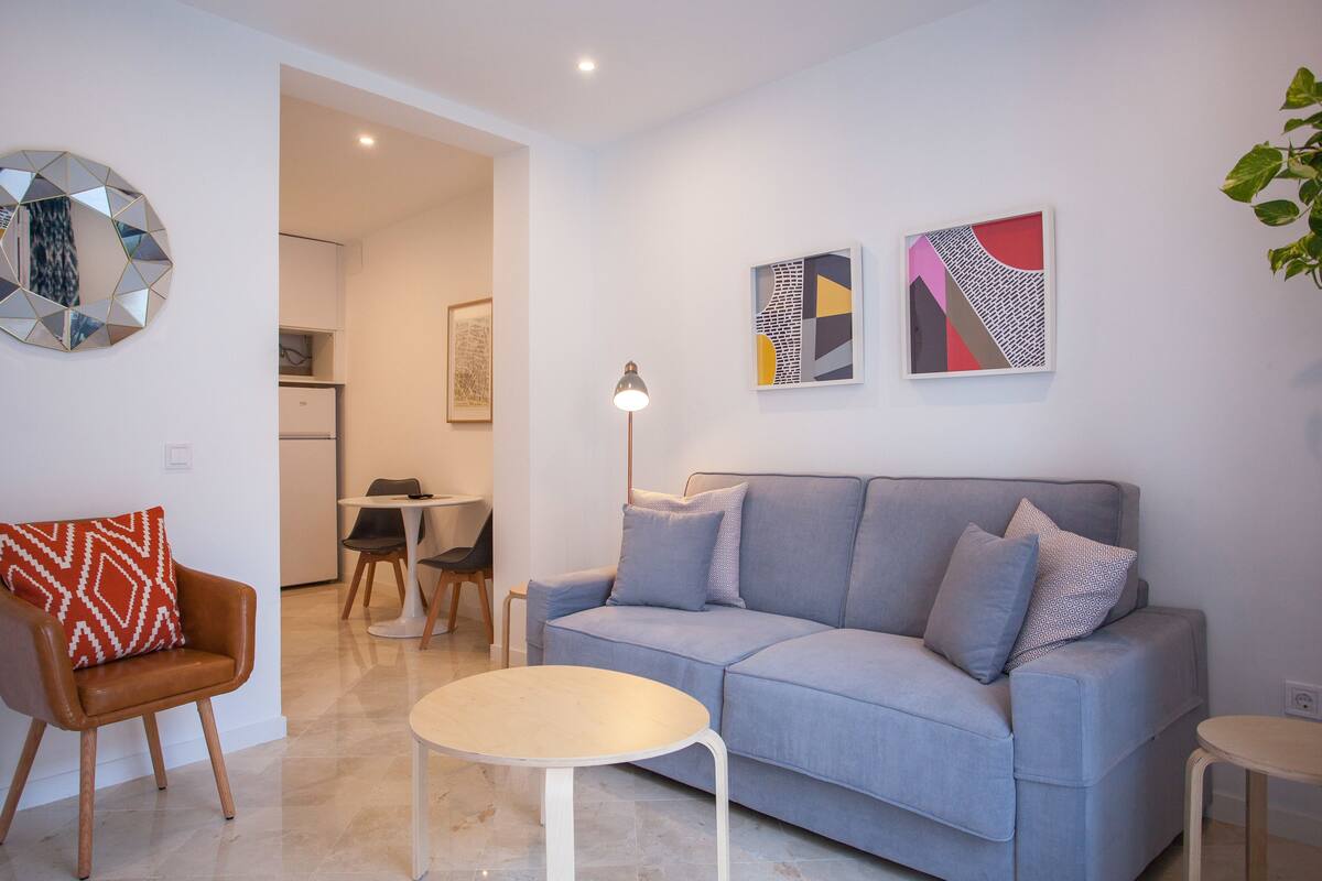 A modern living room with a grey sofa, a round coffee table, and a brown armchair with a patterned cushion. There are two abstract paintings on the wall and a floor lamp.