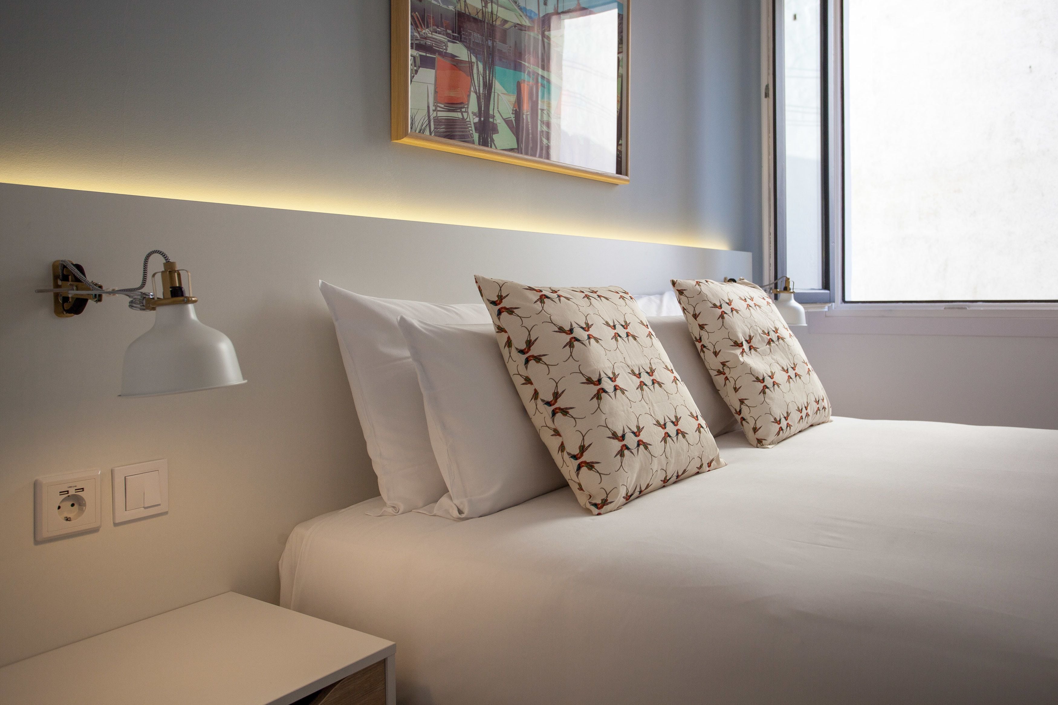 Un lit bien fait, avec une literie blanche et deux coussins à motifs, une lampe de chevet et un luminaire fixé au mur.