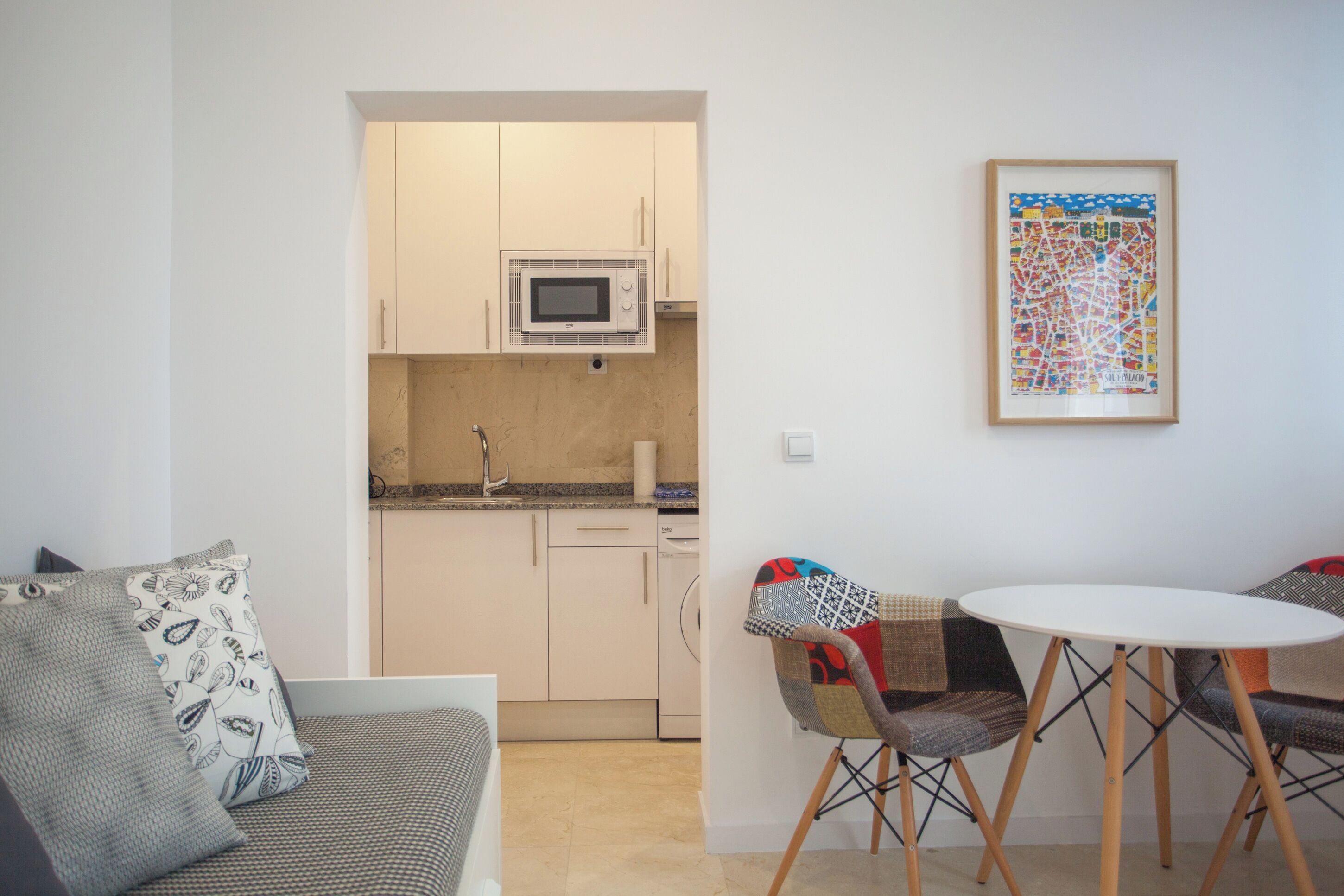 Un espace de vie compact comprenant un coin cuisine, un coin repas avec une table ronde et des chaises, et un canapé agrémenté de coussins à motifs.
