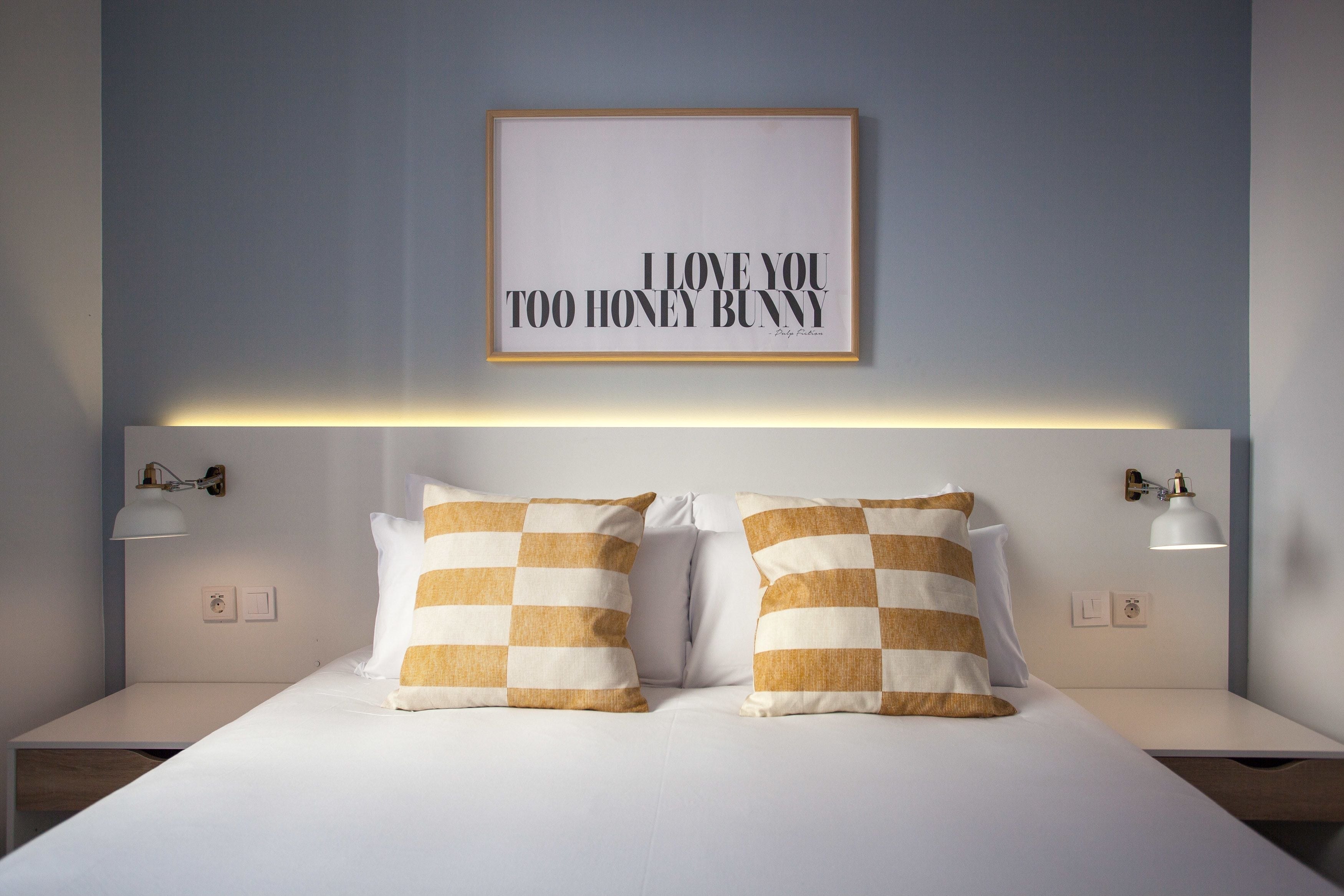 Cama bem arrumada com duas almofadas, um quadro na parede e luminárias de cabeceira.