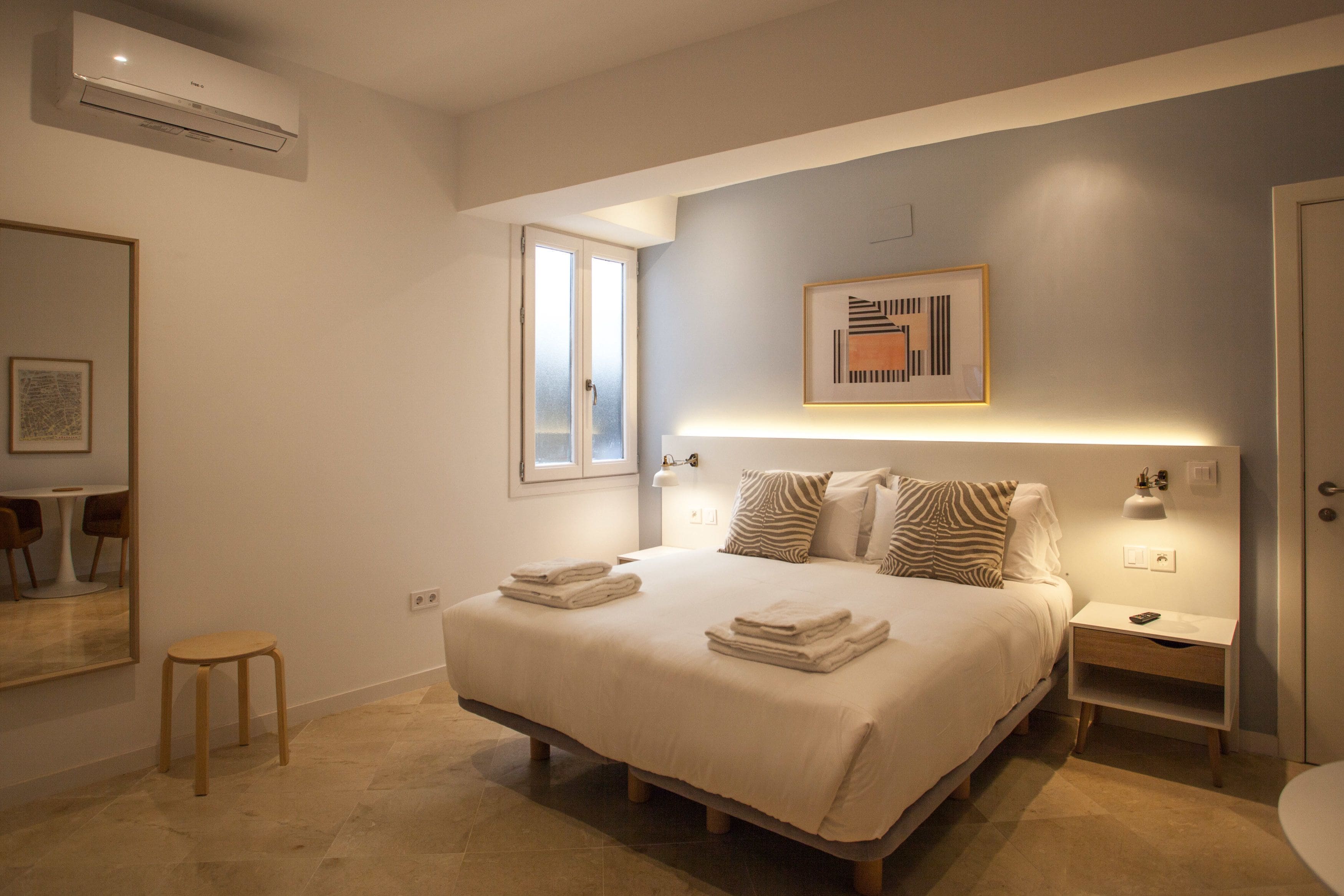 Un dormitorio moderno con una cama grande, mesitas de noche, un taburete pequeño, un ventanal y una obra de arte abstracta enmarcada en la pared.