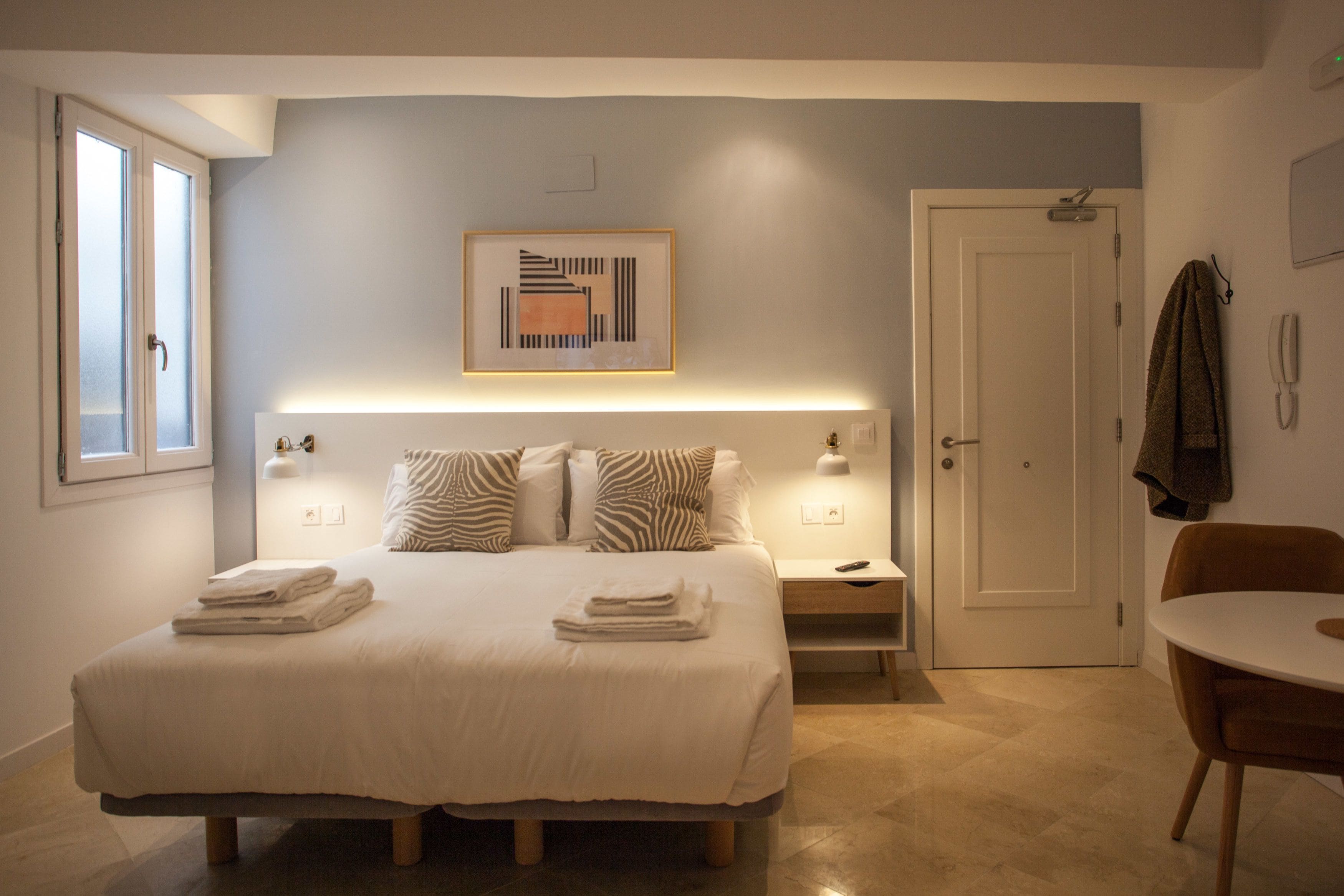 A hotel room with a bed, bedside tables, a chair, a door, and a painting on the wall.