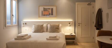 A hotel room with a bed, bedside tables, a chair, a door, and a painting on the wall.