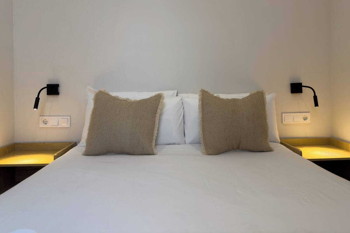 A neatly made bed with two pillows, two bedside tables, and two wall-mounted lamps.