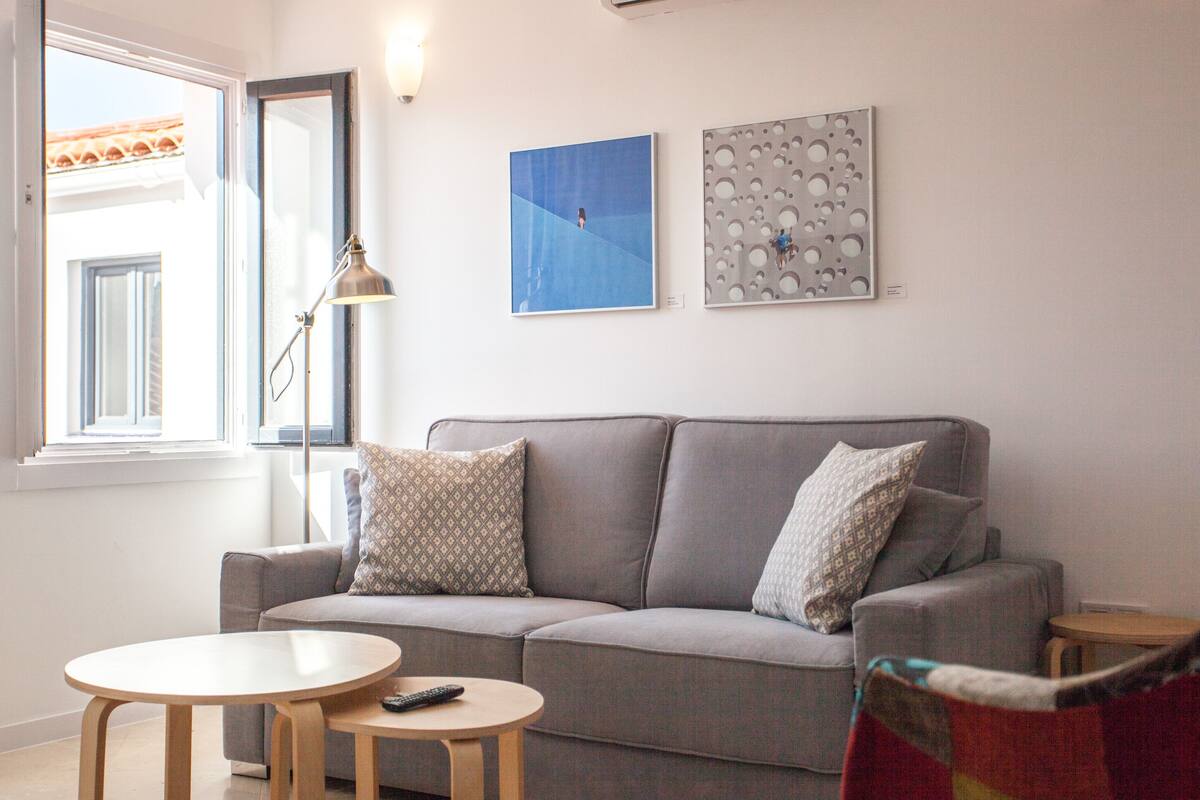 A living room with a grey sofa, two patterned pillows, a round wooden table, and a remote control on it.