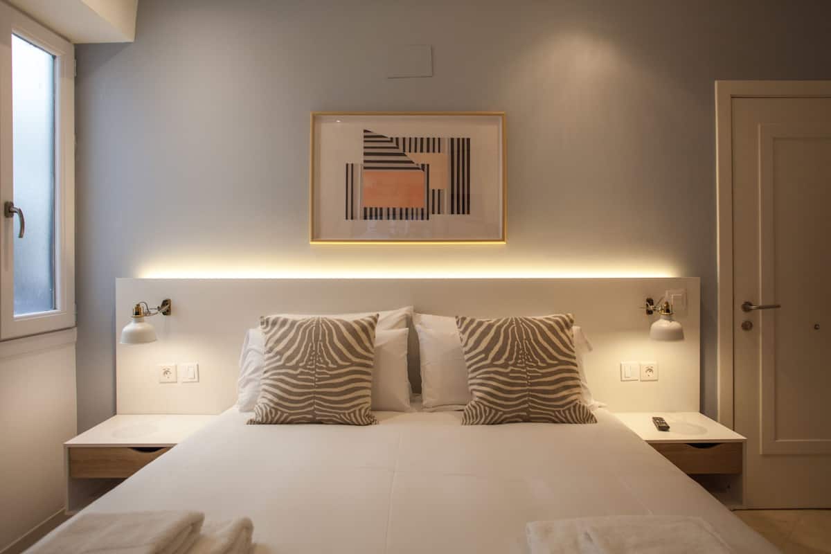 A modern bedroom with a large bed, zebra-striped pillows, bedside lamps, and a framed abstract artwork on the wall.