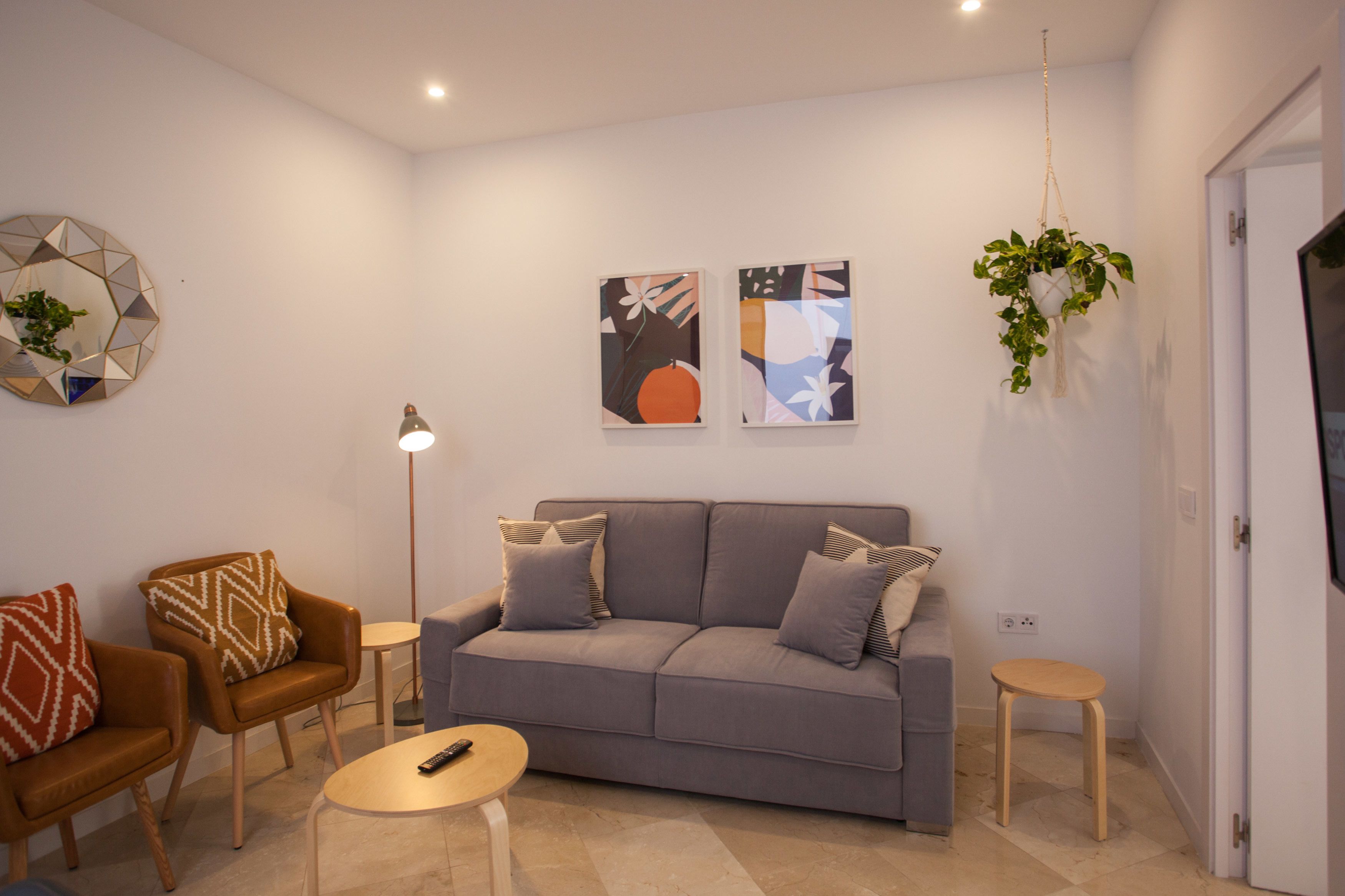 A living room with a grey sofa, two armchairs, a coffee table, and a floor lamp. There are two wall-mounted paintings and a hanging plant.