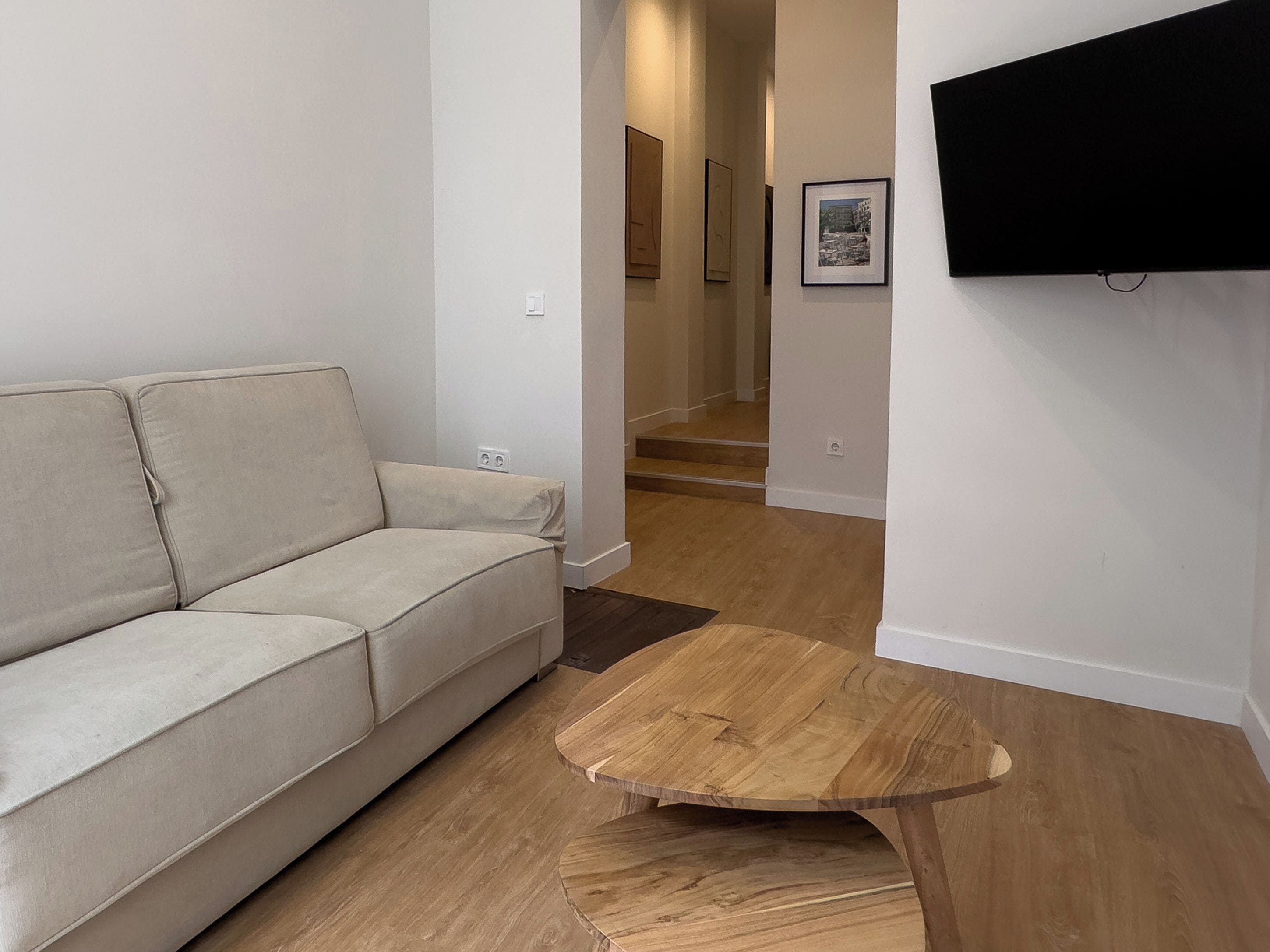 A living room with a beige sofa, a wooden coffee table, and a flat-screen TV mounted on the wall.