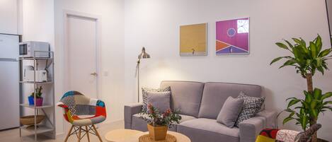 Una sala de estar con un sofá gris, un sillón colorido, una mesita con una planta y una lámpara de pie.