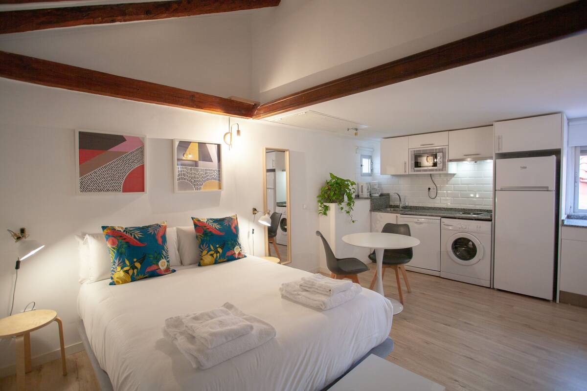 A modern bedroom with a large bed, a small desk, and a kitchenette with a washing machine.