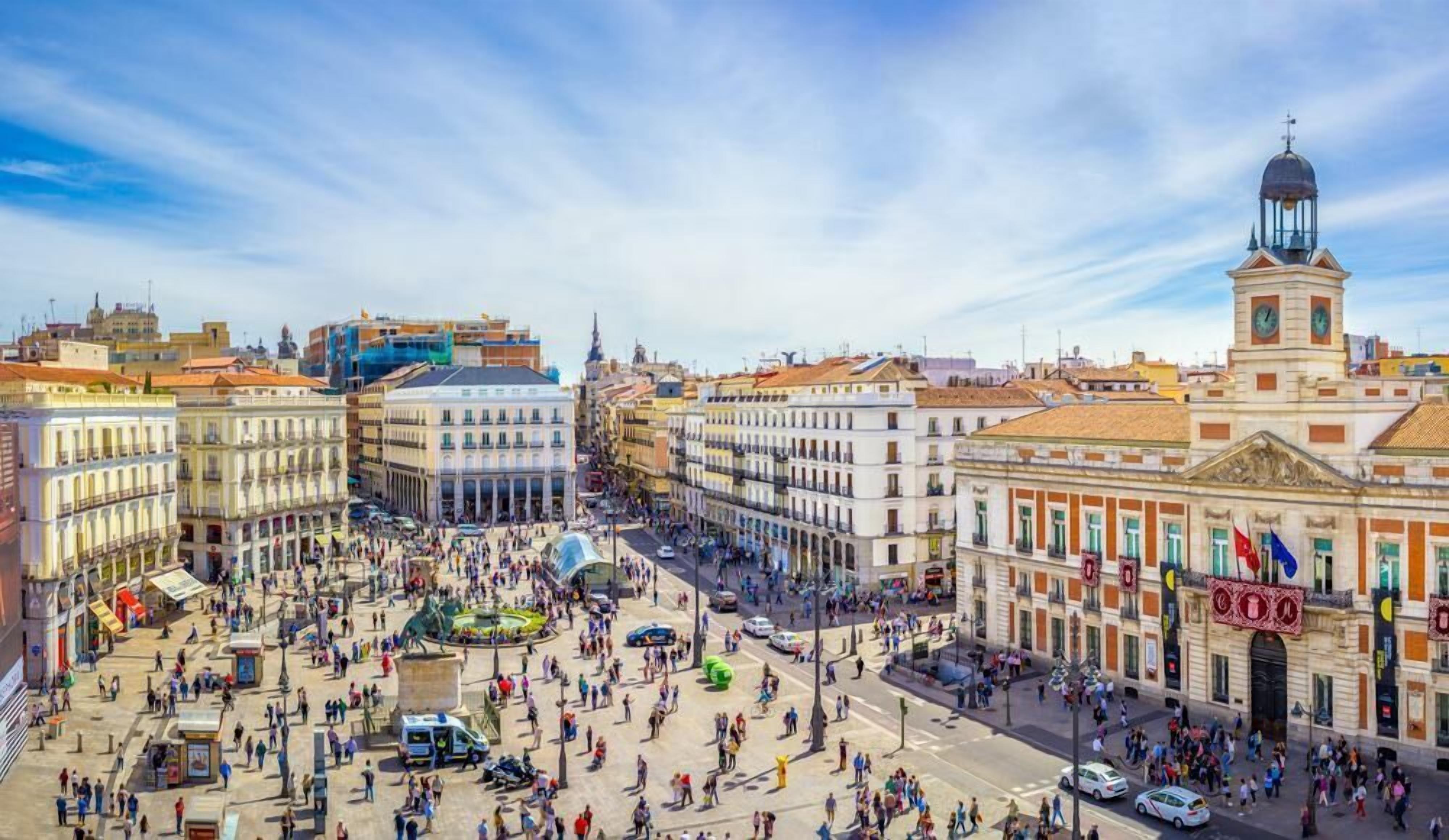 A bustling square with historic buildings, a prominent clock tower, and a central fountain.