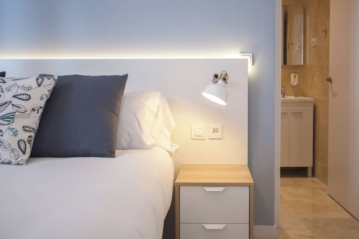 A bedroom with a bed, bedside lamp, nightstand, and a bathroom visible in the background.