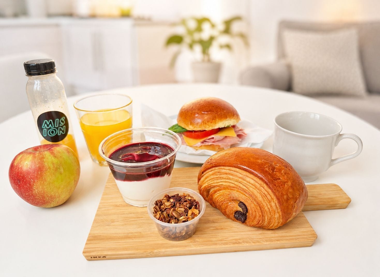 Un desayuno con un croissant, manzana, jugo, yogur y un sándwich sobre una tabla de madera.