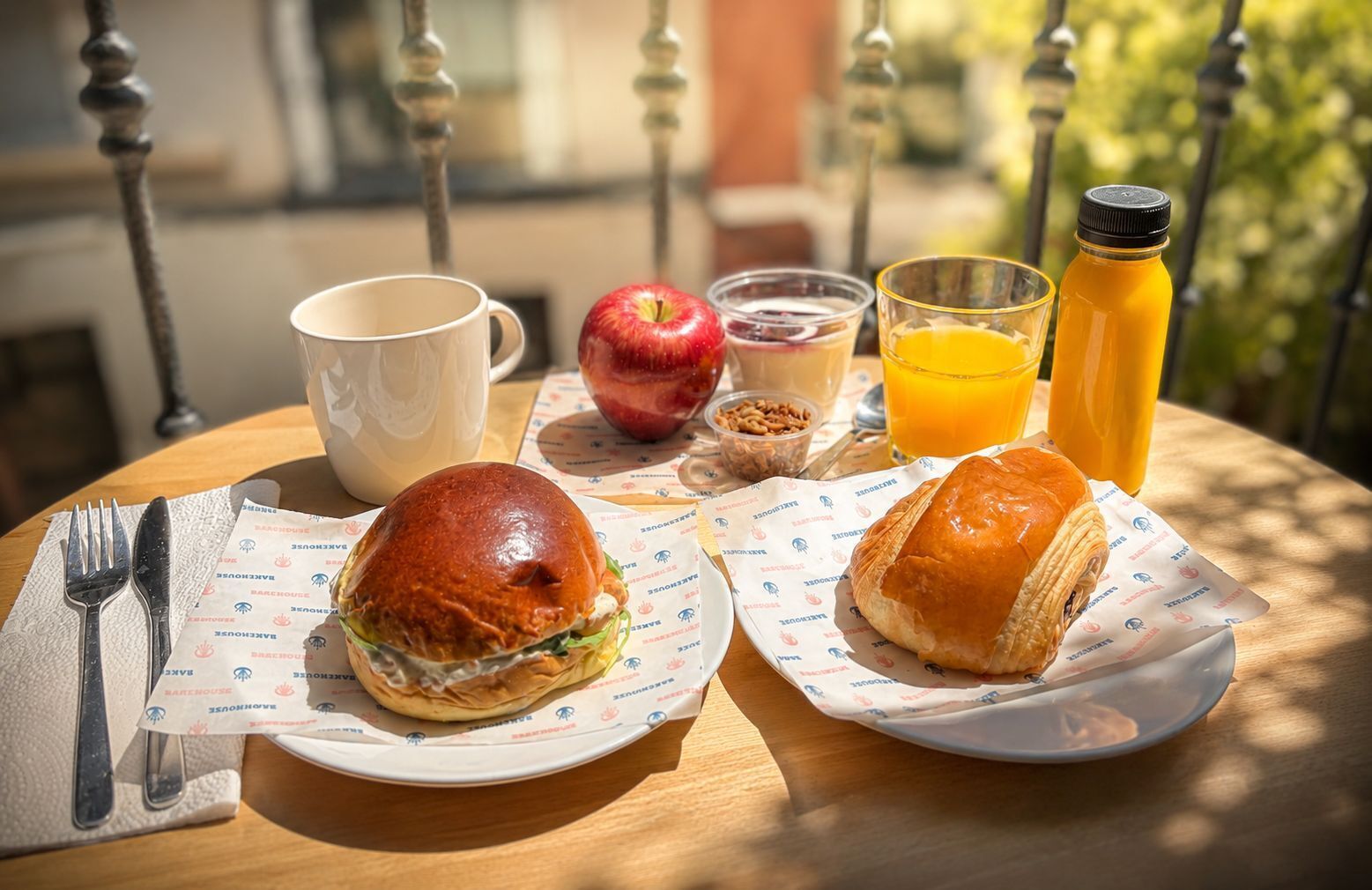 Une table avec un sandwich, un croissant, une tasse, un verre de jus, une bouteille de jus et un petit récipient contenant des noix.