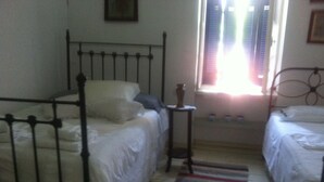 A small, dimly lit room with two beds, a small table, and a window with blinds. - Persefonis view house (Skopelos)