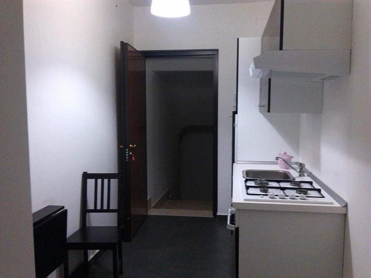 A narrow hallway leading to a door, a small kitchen area with a gas stove and sink, and a single chair.
