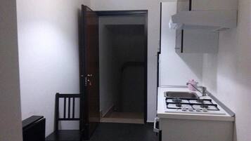 A narrow hallway leading to a door, a small kitchen area with a gas stove and sink, and a single chair.