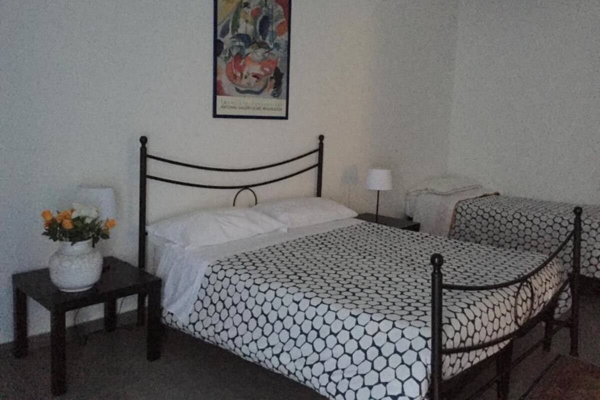 A room with a bed, a nightstand with a vase of flowers, a lamp, and a painting on the wall.