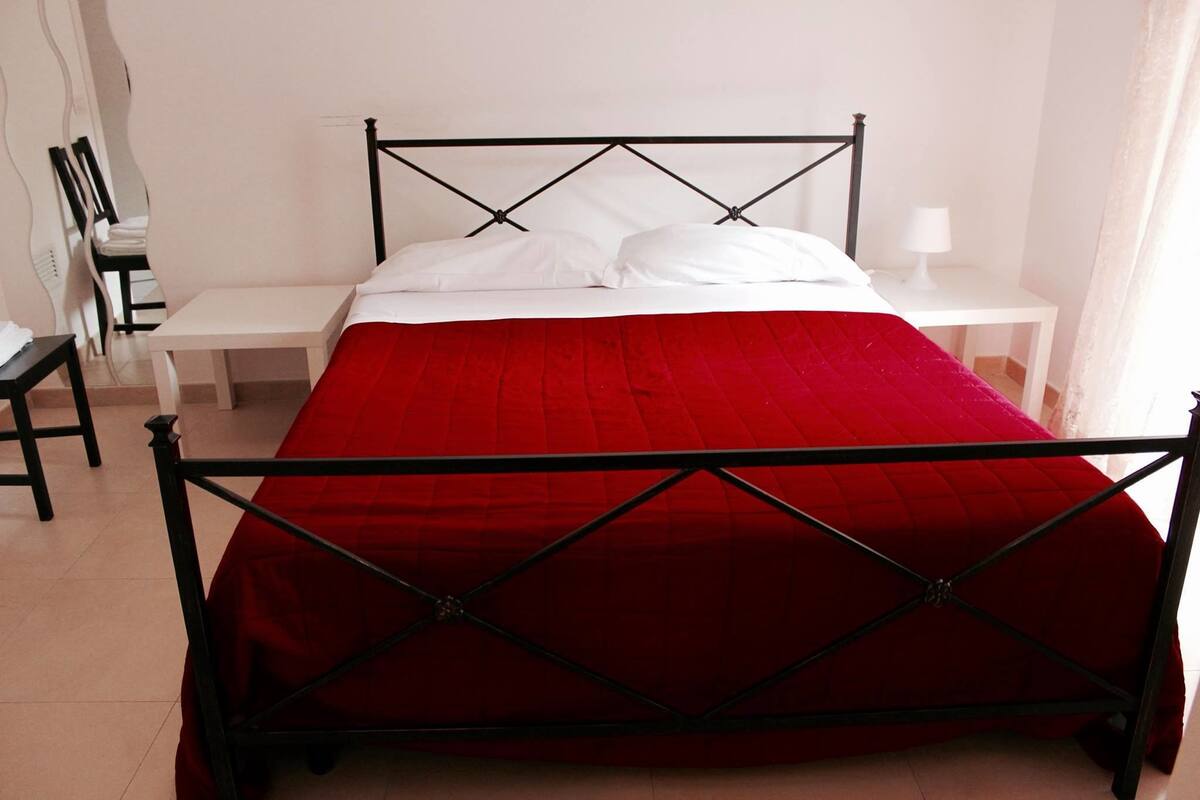 A bed with a red bedspread and white pillows, a small bedside table with a lamp, and a chair in a room with white walls and a tiled floor.