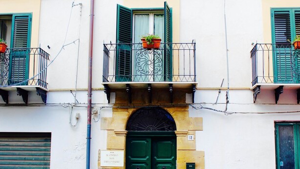 Un edificio con puerta verde, un balcón con macetas y un cartel sobre la puerta.