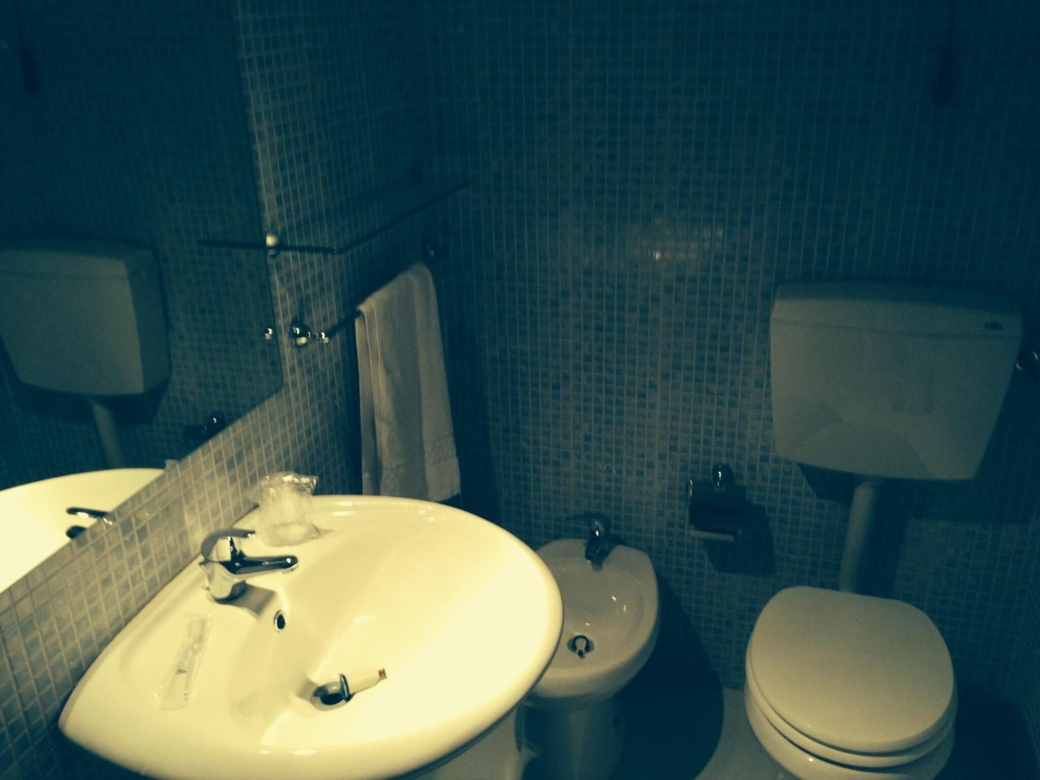 Une salle de bain équipée d’un lavabo, de toilettes et d’un bidet. Les murs sont recouverts de carreaux, et un miroir est placé au-dessus du lavabo.