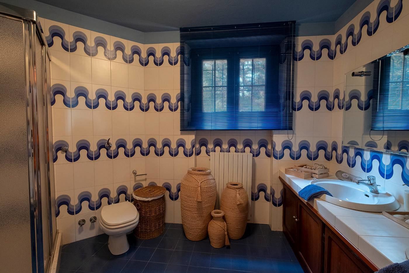 Ein Badezimmer mit gefliestem Rückspritzschutz, Toilette, Waschbecken und einem Fenster mit Jalousien.