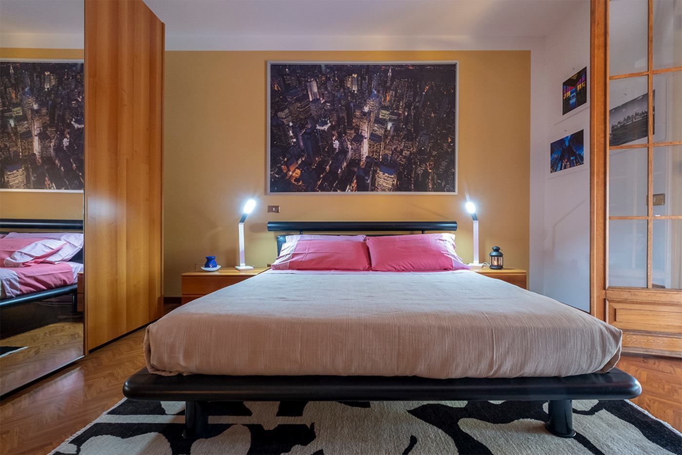 A modern bedroom with a large bed, two bedside tables with lamps, a cityscape painting on the wall, and a wooden wardrobe.