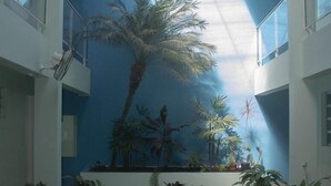 A spacious atrium with a high ceiling, a skylight, and a central area featuring a large potted plant. There are wicker chairs and a small table in the foreground. - Hotel Porto Fino (Pontal do Parana)