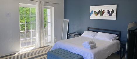 Habitació amb un llit, un pouf blau, una tauleta de nit, un quadre i vista a verdura a través d'una porta corredissa.