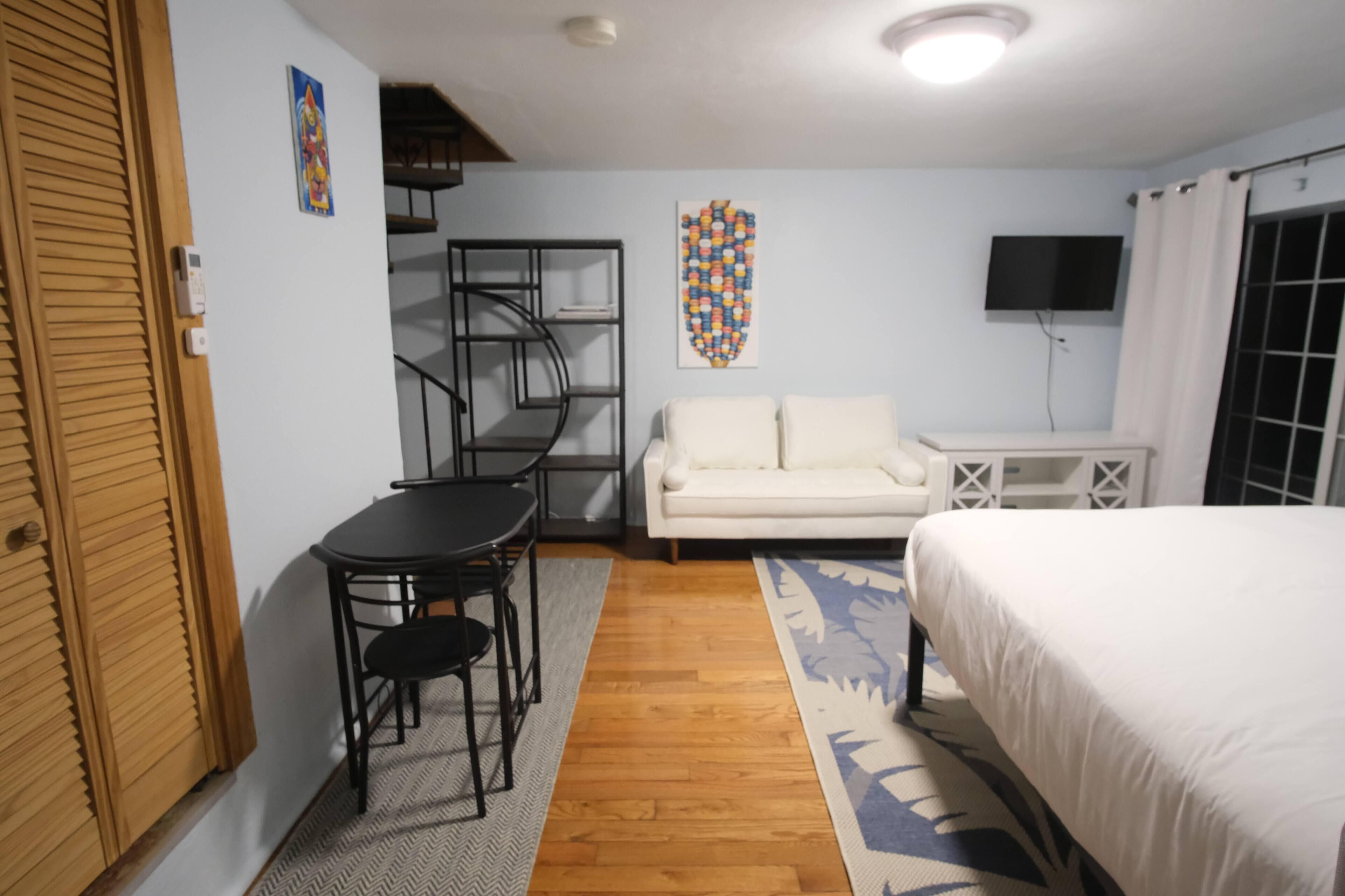 A room with a bed, a white sofa, a small table with two chairs, a TV, and a wall art piece.